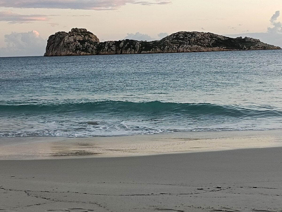 A serene coastal view captures a rocky island in the distance, framed by calm turquoise waters. Gentle waves lap at a sandy beach, which reflects soft hues from a setting sun above, creating a peaceful atmosphere.