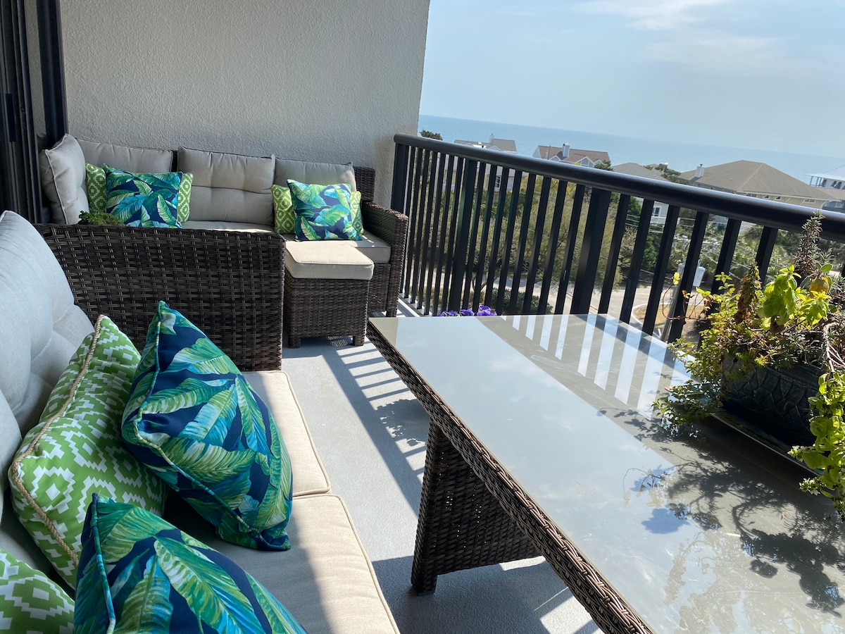 A balcony seating area is furnished with comfortable wicker couches and a glass-top table. Brightly patterned cushions adorn the seating. The expansive view of the ocean is visible in the background, framed by a railing and gently swaying plants.