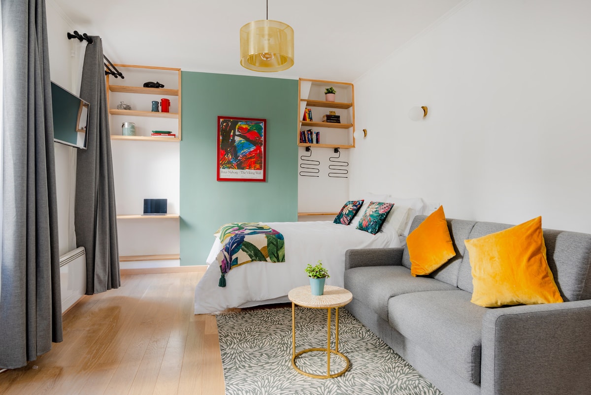 A cozy studio features a comfortable bed adorned with colorful accents and a sofa with decorative pillows. A small table sits on a patterned rug, and shelves hold books and decorative items. Light curtains frame the window, allowing natural light to brighten the space.