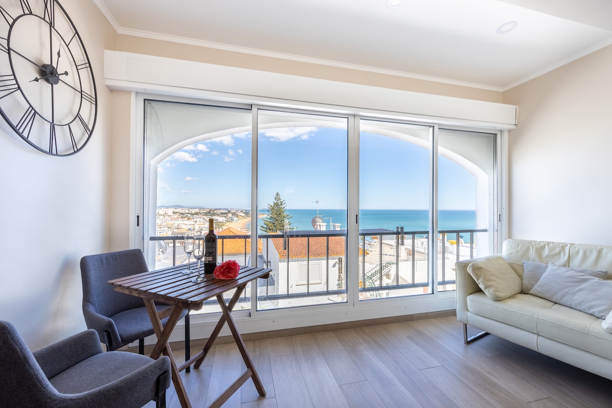 A bright sitting area is showcased with large glass doors offering expansive views of the sea and surrounding landscape. A small table with two chairs sits next to a light-colored sofa. Natural light fills the room, enhancing the calm environment.