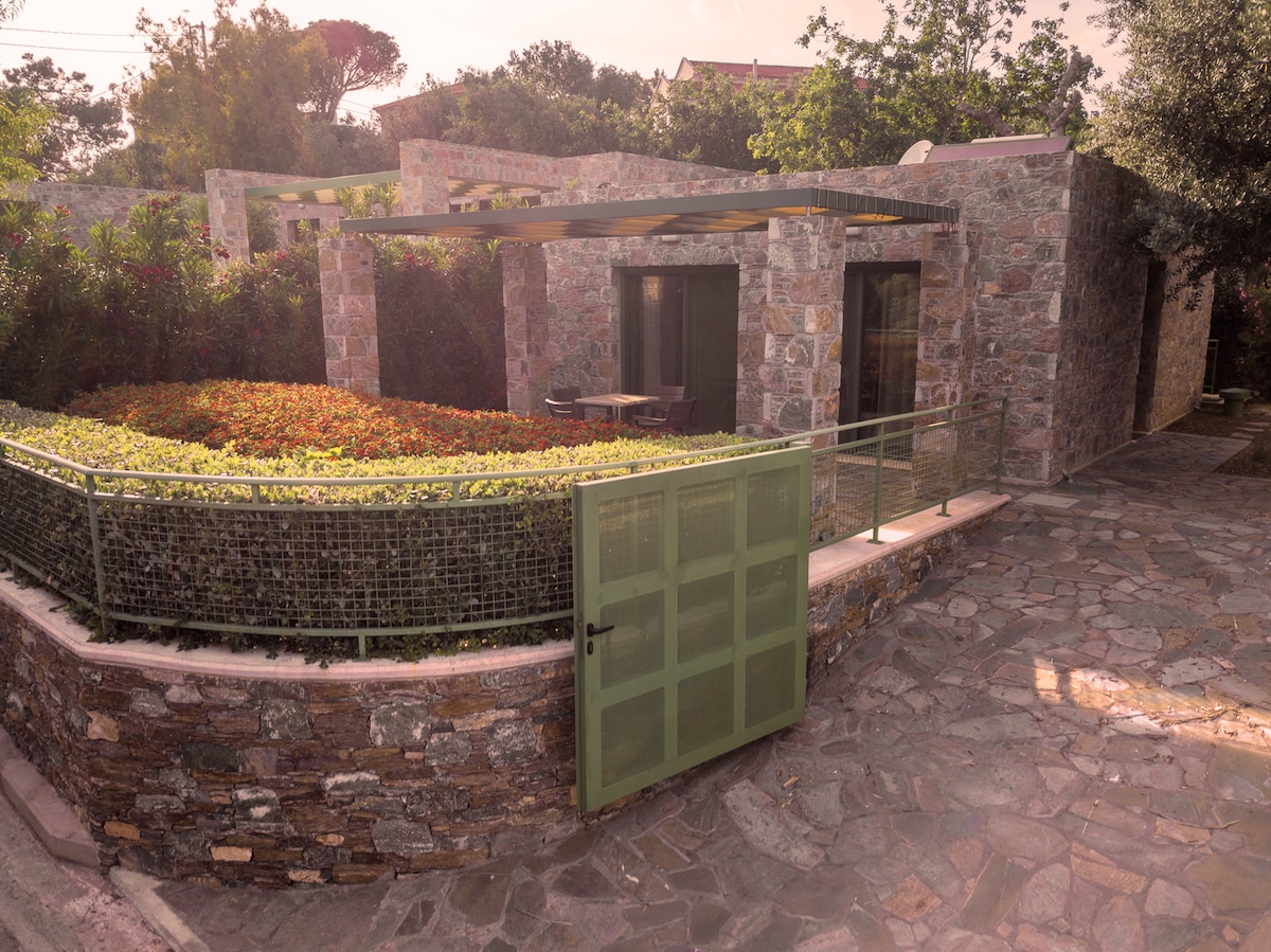 The exterior of the Stone Studio is highlighted by natural stone walls, encompassing a garden area with lush greenery and vibrant plants. A simple gate leads to a stone pathway, while a pergola provides shade over an outdoor seating area.