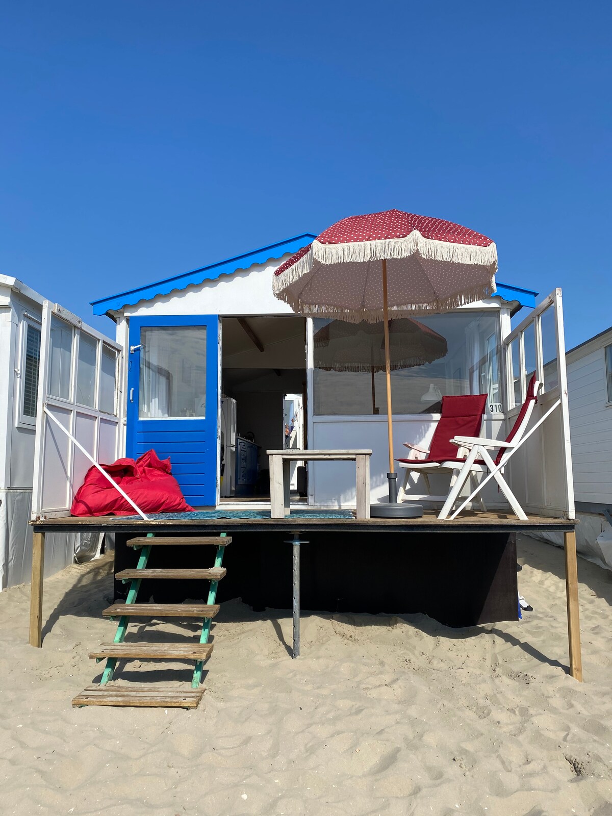 A beach house is depicted with a bright blue door and a large red and white umbrella shading a seating area. Two chairs and a small table are positioned on a deck overlooking the sandy beach, while steps lead down to the shore.