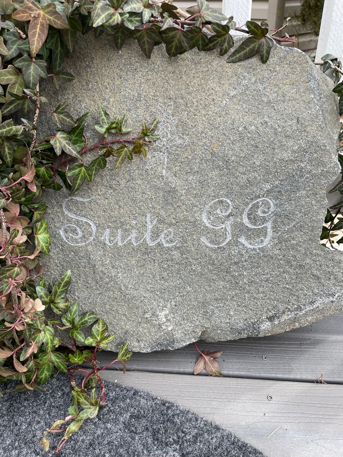 A natural stone engraved with 'Suite GG' is surrounded by trailing ivy, positioned on a wooden surface. The stone adds an organic touch, serving as a welcoming feature for guests arriving at the apartment.