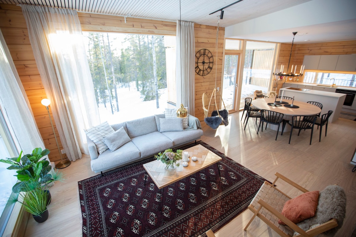 A welcoming living area features a spacious sofa and a glass coffee table resting on a decorative area rug. Large windows allow natural light to illuminate the room, with a view of the snowy outdoors. Dining space with modern chairs and swing seats is visible in the background.