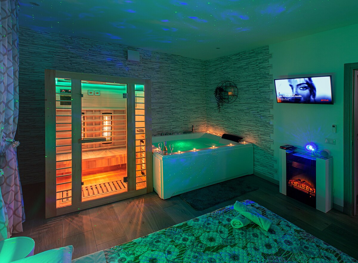 A tranquil room is illuminated with soft green light, featuring a spacious Jacuzzi tub with candles around it. A sauna is visible through wooden slats, and a wall-mounted television offers entertainment. The decor includes textural stone walls and a cozy electric fireplace enhances the ambiance.