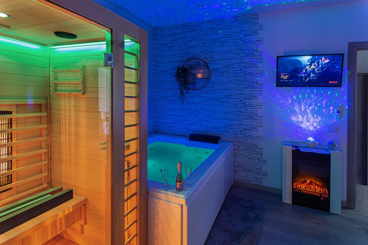 A wellness space features a sauna with a wooden bench and ambient lighting. Adjacent, a jacuzzi bath is visible, complemented by a wall-mounted television. Decorative elements and soft lighting enhance the atmosphere, creating a serene retreat for relaxation.