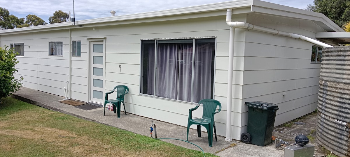 A view of the exterior of the guest apartment showcases a single-story, white-cladded building. Two green chairs are positioned on a small concrete patio in front of a large window. A trash bin and garden area are visible alongside the structure, creating a tidy and accessible environment.