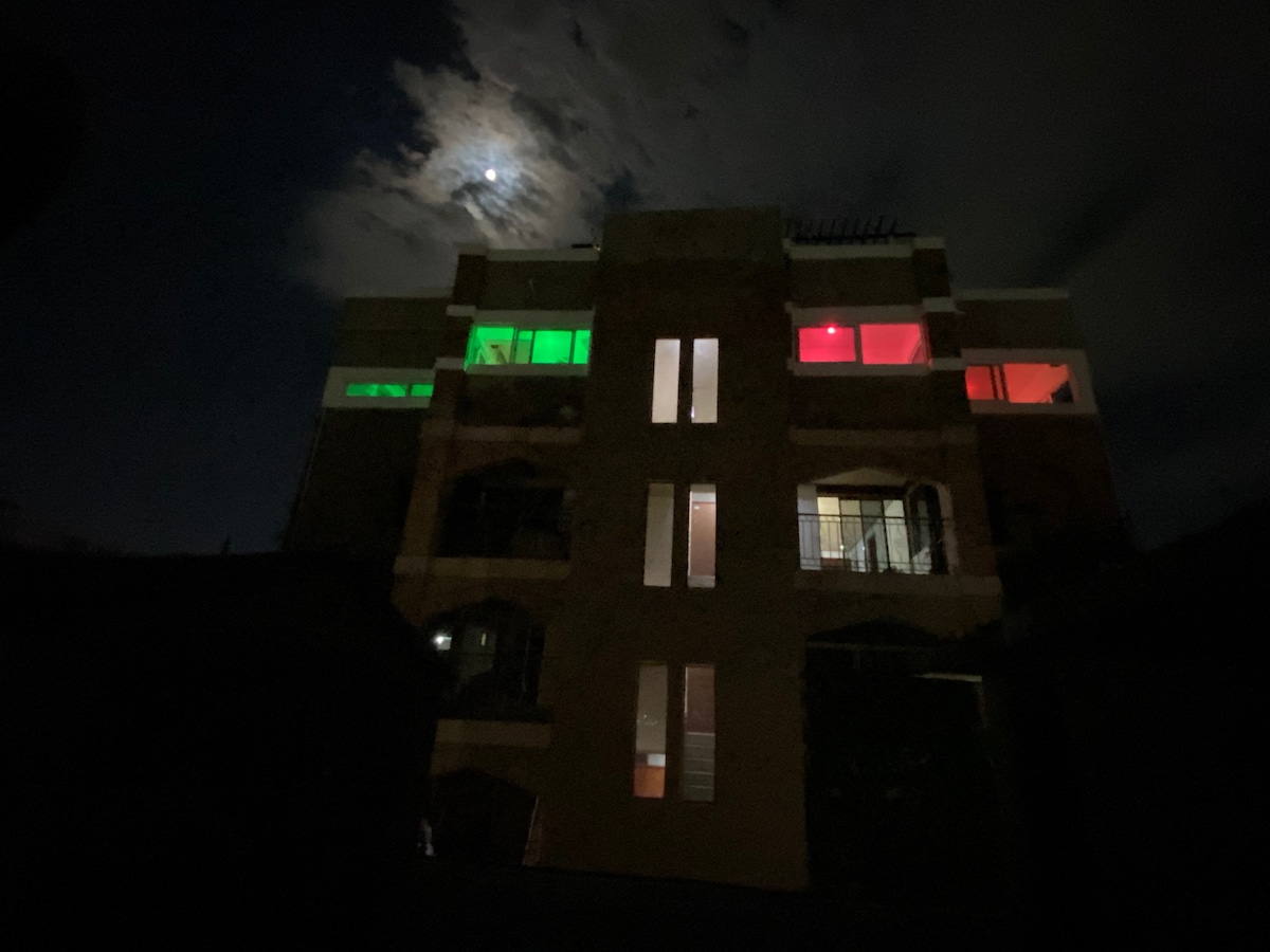 An exterior view of a multi-story building is captured at night, illuminated with red and green lights. The design includes large windows, with some rooms brightly lit, while the moonlight adds a soft glow to the scene.
