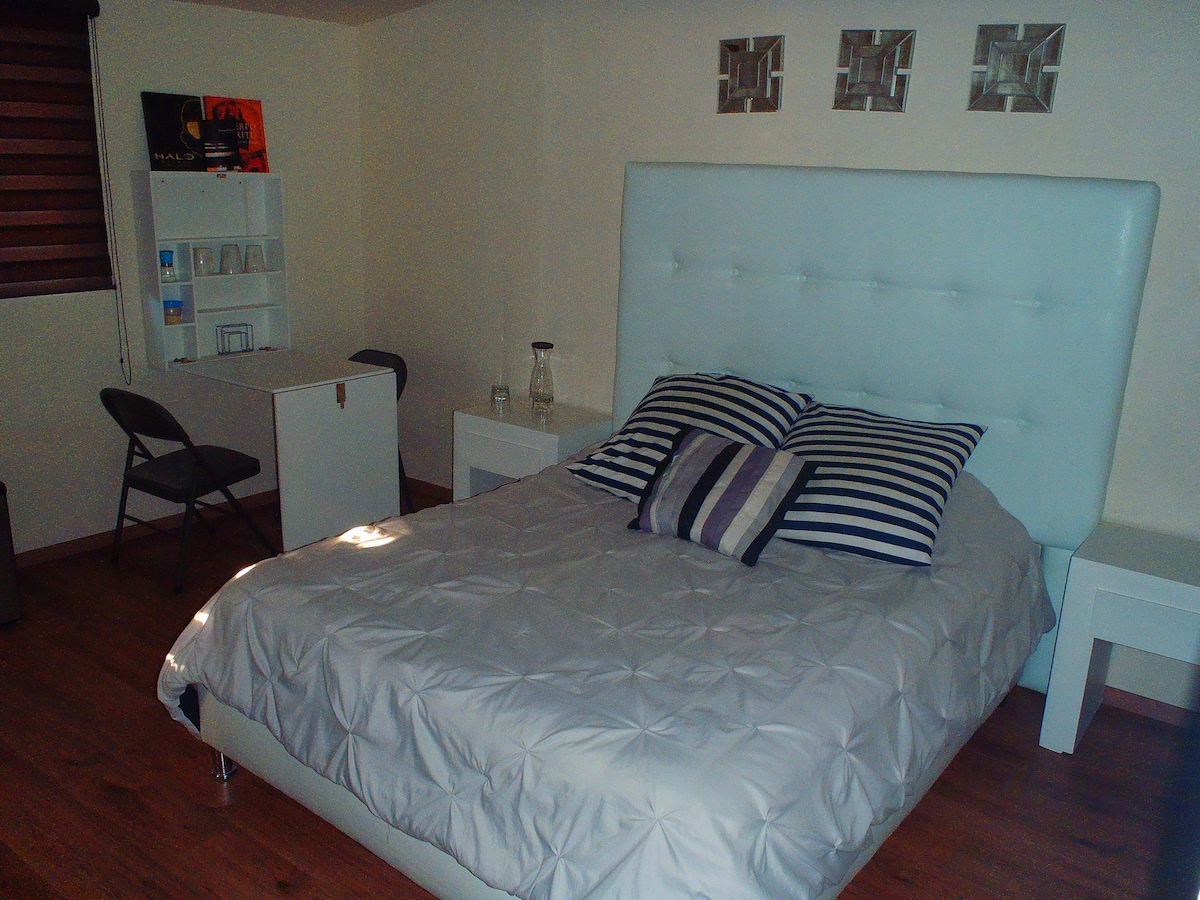 A cozy sleeping area features a large, tufted white bed with striped pillows. Nearby, a small desk with two folding chairs is positioned against the wall. Natural light enters through a window with wooden blinds, illuminating the warm wooden floor.