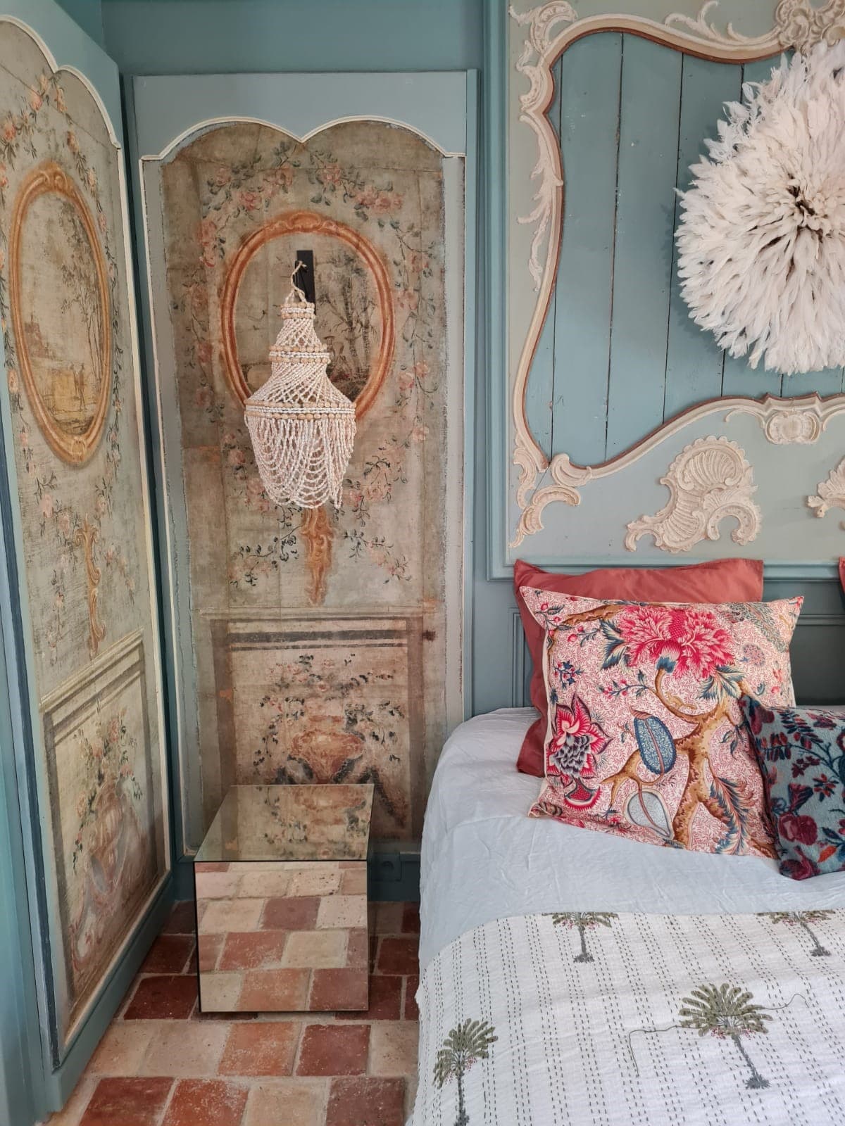 A cozy corner of the room features an intricately designed floral panel on the wall, complemented by a stylistic woven hanging piece. A comfortable bed adorned with colorful pillows is visible, alongside a small glass table resting on tiled flooring.