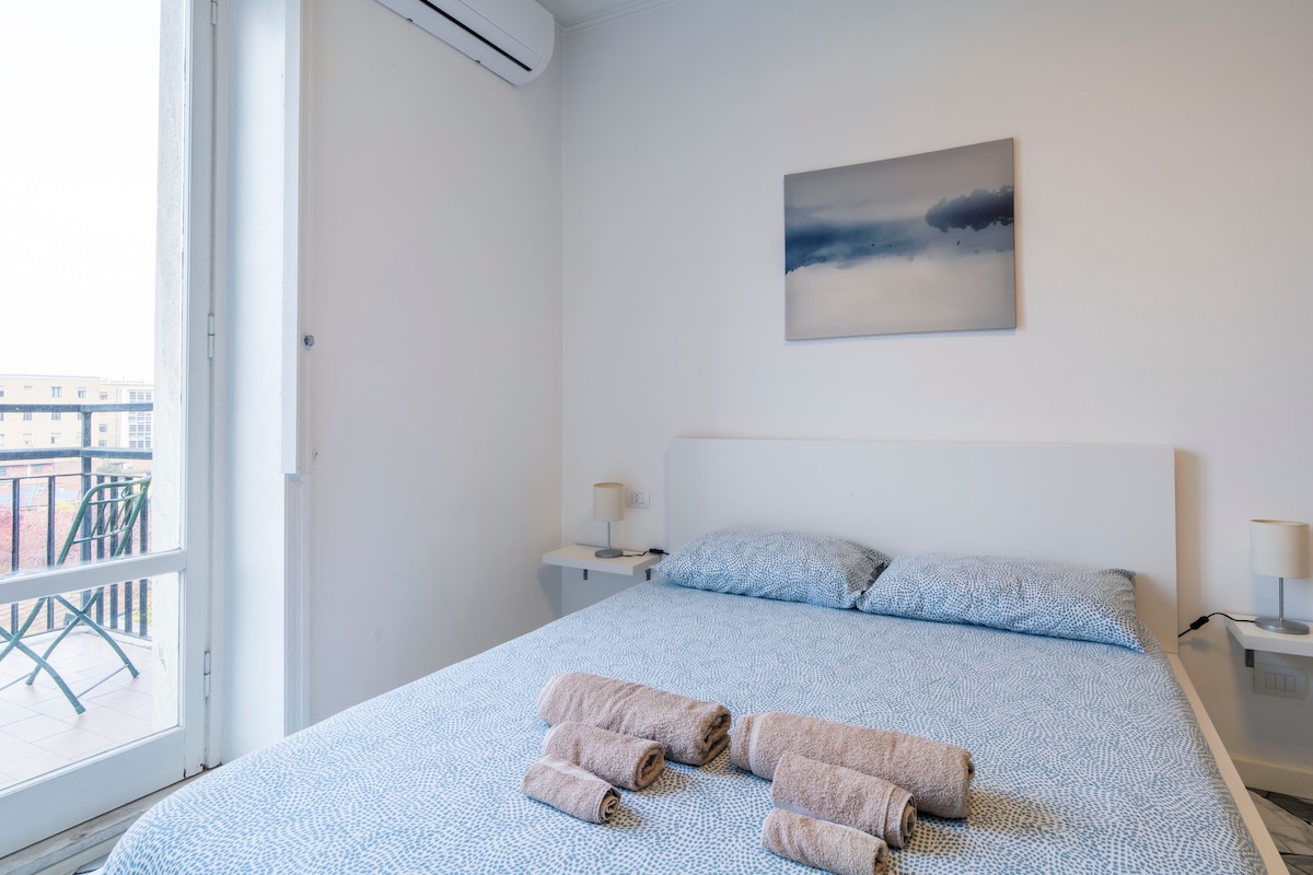 A light-filled bedroom features a neatly made spacious bed dressed in patterned linens. Clean towels are arranged on the bed, and a simple bedside table holds stylish lamps. A window provides a view of the outside, enhancing the airy feel of the room.
