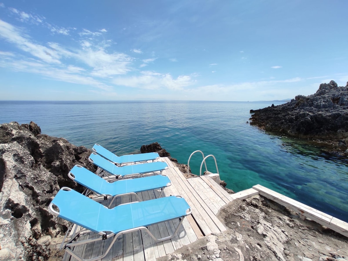 A wooden deck extends over calm, clear waters, featuring several lounge chairs arranged in a row. Rocky shorelines frame the serene setting, while the horizon showcases a blend of blue skies and distant land. The gentle waves create a tranquil atmosphere.