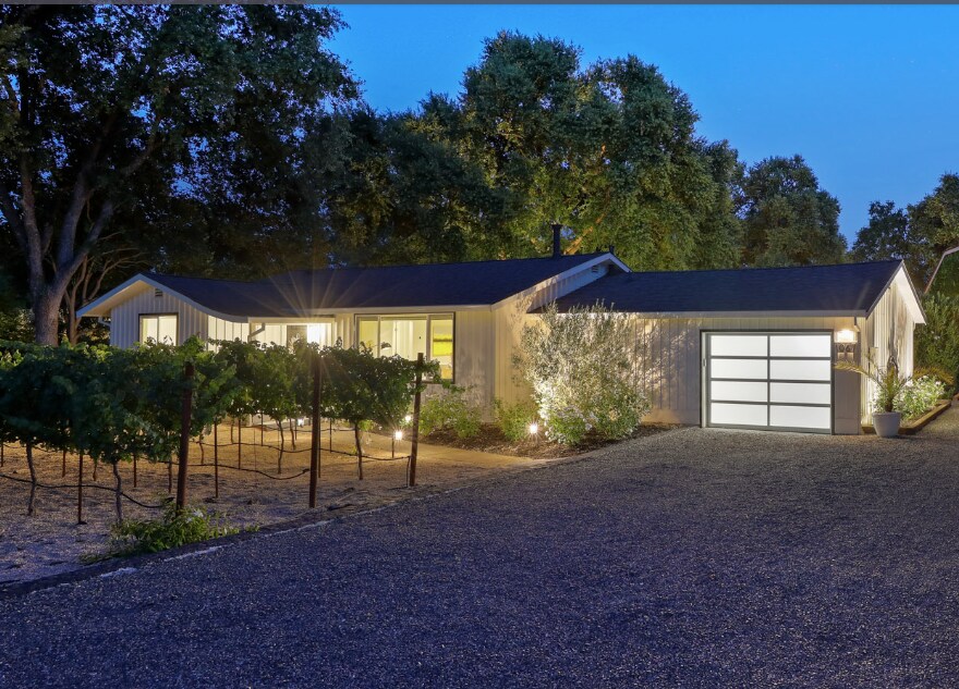The exterior of the updated farmhouse is illuminated at dusk, featuring modern architecture with large windows. A well-maintained gravel driveway leads to a sleek garage door. Lush landscaping includes a row of vines in the foreground and mature trees providing a natural backdrop.