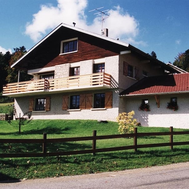 The exterior of a chalet-style house is shown, featuring a sloped roof and a large balcony. The facade displays a combination of wood and stone materials. A lush green lawn surrounds the property, with a wooden fence defining the boundary. Blue skies and fluffy clouds provide a serene backdrop.