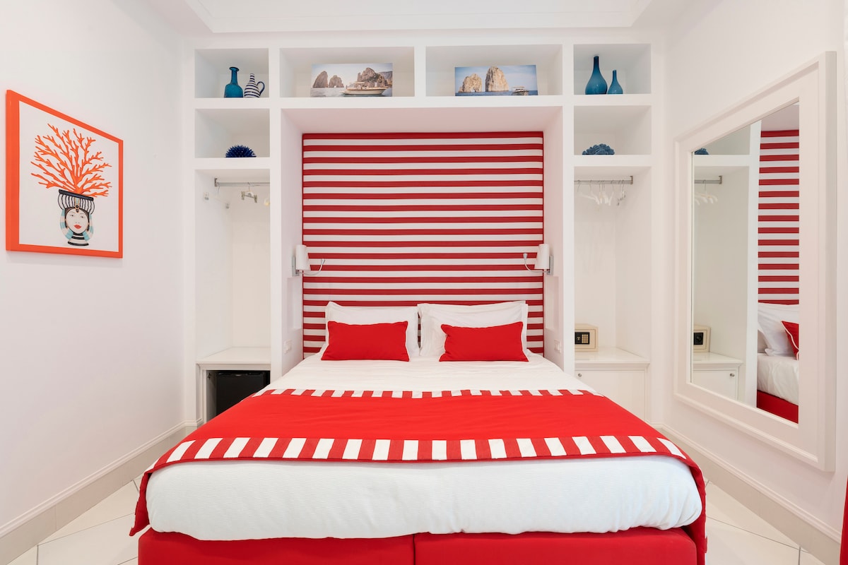 A modern bedroom features a spacious bed with red and white striped linens. A red accent wall is complemented by white shelving displaying decorative items. Natural light is provided by a nearby mirror reflecting the room's vibrant colors.