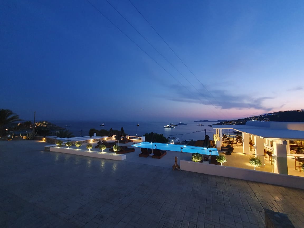 The outdoor area is illuminated with soft lighting, highlighting the infinity pool that stretches along the patio. Lounge chairs are positioned beside the pool, offering views of the sea and sunset. The horizon features distant boats, contributing to the serene ambiance.