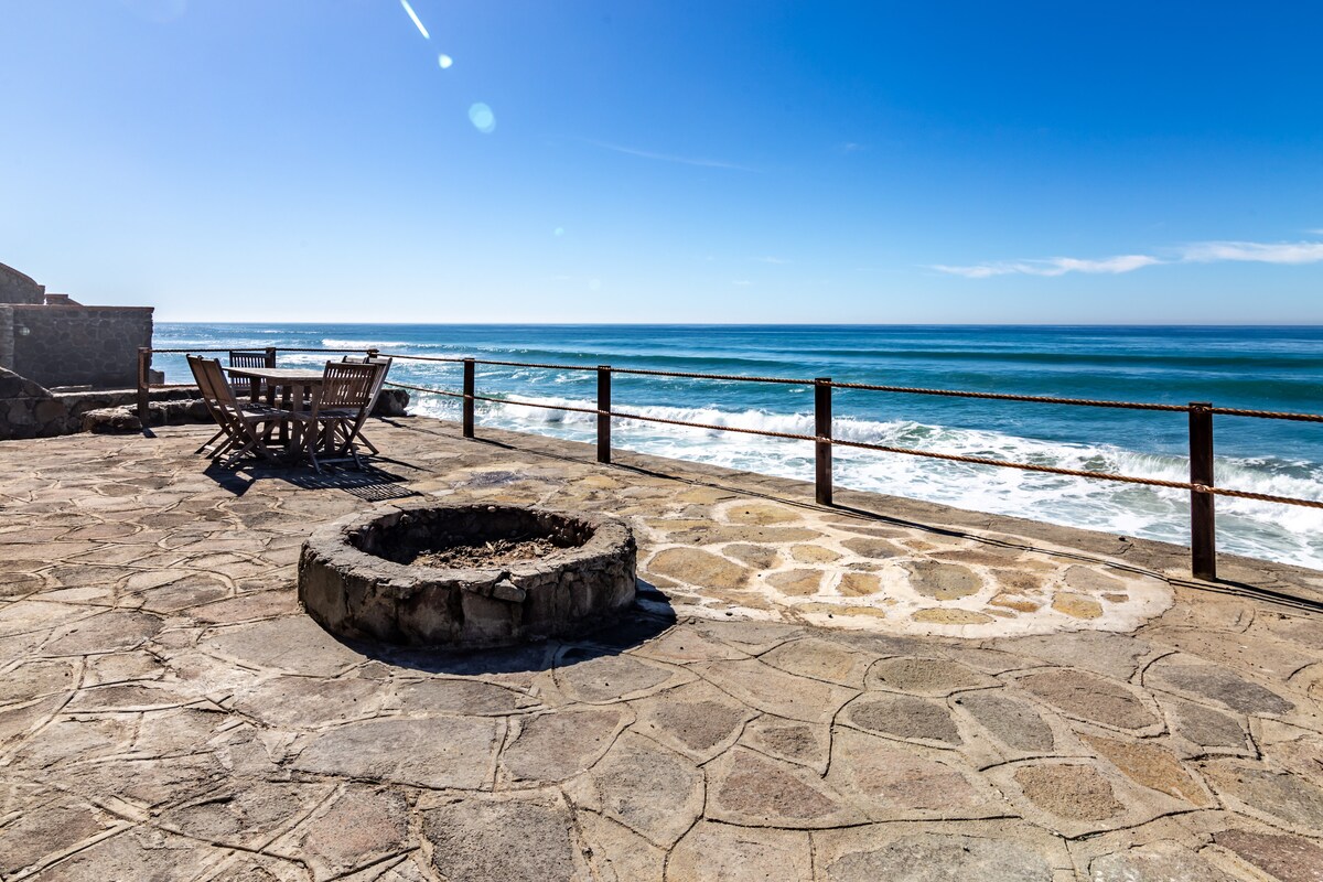 An oceanfront patio features a natural stone surface with a circular fire pit at the center. A rustic wooden dining table with chairs is positioned nearby, offering panoramic views of the ocean waves and clear blue sky, enclosed by a low railing.