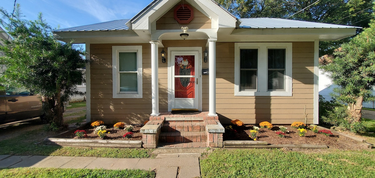 The charming entrance features a red door framed by white columns, flanked by large windows. A well-maintained flower bed with colorful flowers complements the exterior. The property is surrounded by a grassy yard, with a walkway leading to the front steps.