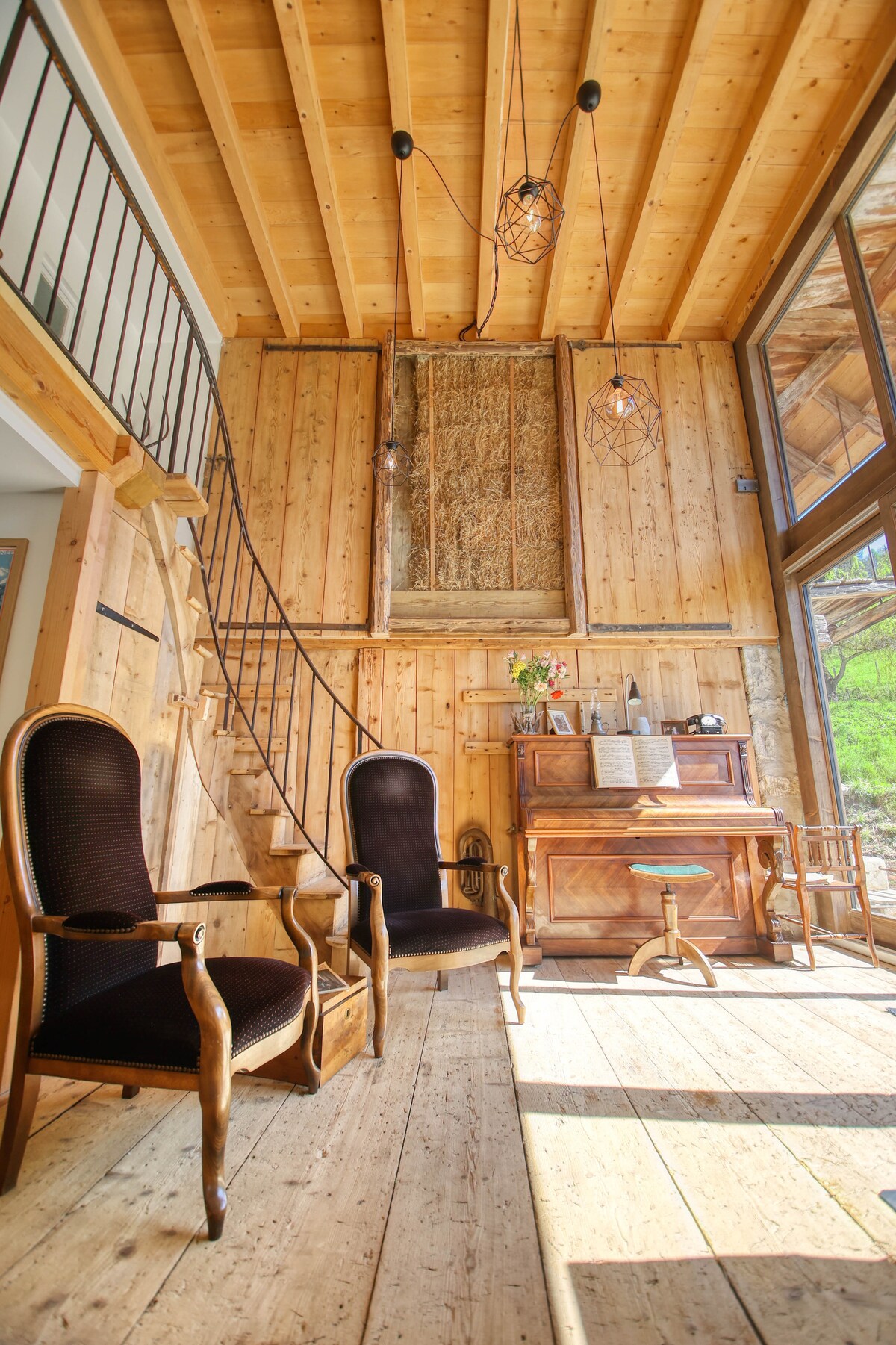A sunlit living area features timber walls and flooring, complemented by two elegant chairs. A wooden piano is positioned beside a large window, offering views of nature. A spiral staircase leads to the upper level, while modern light fixtures add a touch of character.
