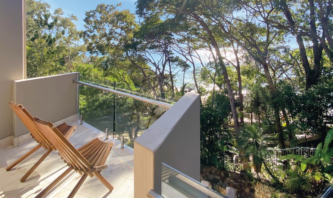 A balcony is shown featuring two wooden lounge chairs facing a serene view of lush green trees and the ocean beyond. The space is illuminated by natural light, enhancing the tranquil atmosphere of the surrounding landscape.