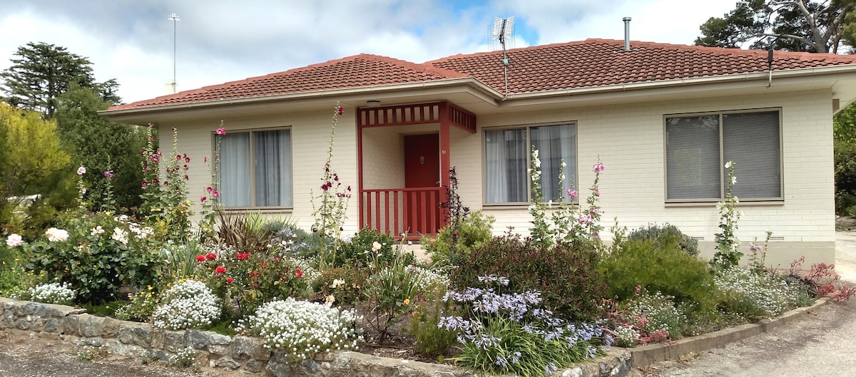 A single-story home is surrounded by well-maintained gardens filled with various flowers. The facade features a red accent on the entrance and large windows that allow natural light. A gravel pathway leads to the front door, enhancing the welcoming appeal of the property.