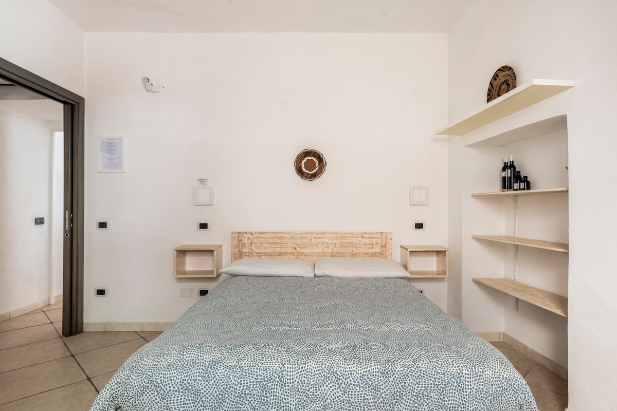A spacious bedroom features a wooden headboard with soft bedding in calming tones. Shelves are positioned above side tables, and a light wall complements the room's neutral decor. Natural light fills the space through a nearby door leading to an additional area.