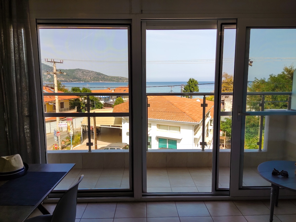 Large glass sliding doors provide a view of the turquoise sea and the nearby evergreen Ipsario Mountain. The balcony is visible, framed by modern railings, while neighboring rooftops and streets create a sense of proximity to the local environment.