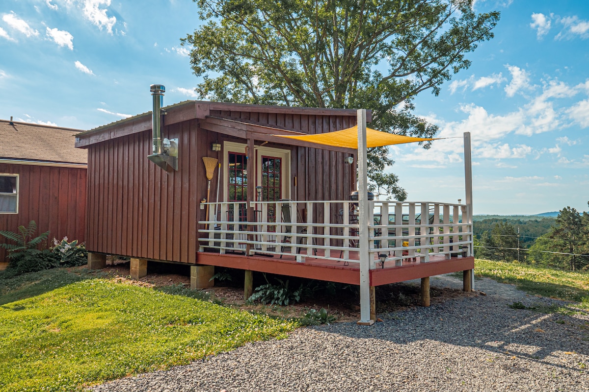 A cabin with a wooden exterior features a covered deck supported by white railings. A chimney protrudes from the roof. Surrounding greenery adds a natural touch, while the expansive landscape in the background can be seen under a clear blue sky.