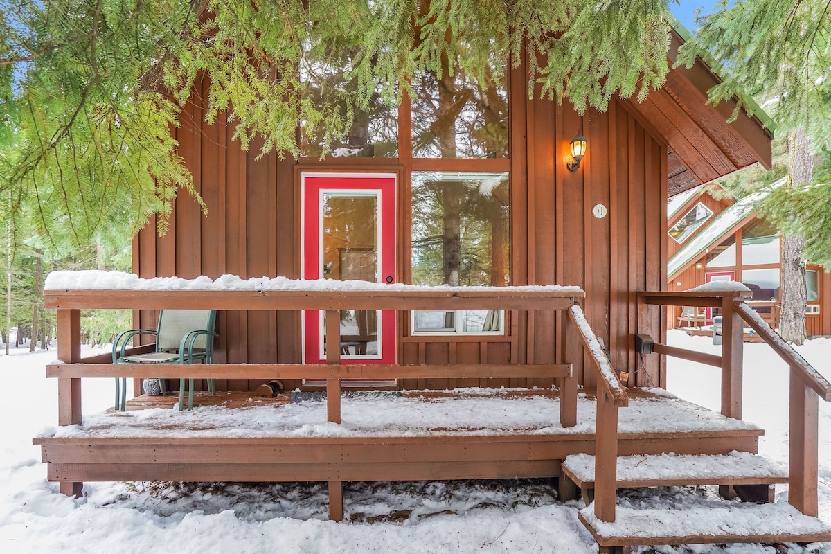 A charming cabin exterior is shown, featuring a wooden facade with a bright red door. A covered porch with a railing offers seating and a few steps leading to the snowy ground, surrounded by tall evergreen trees.