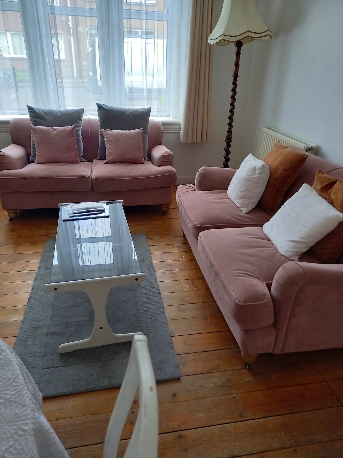 A comfortable seating area features two soft pink sofas adorned with cushions in muted tones. A light wooden coffee table is positioned in the center, resting on a neutral area rug. Natural light filters through large windows, illuminating the warm wooden flooring.