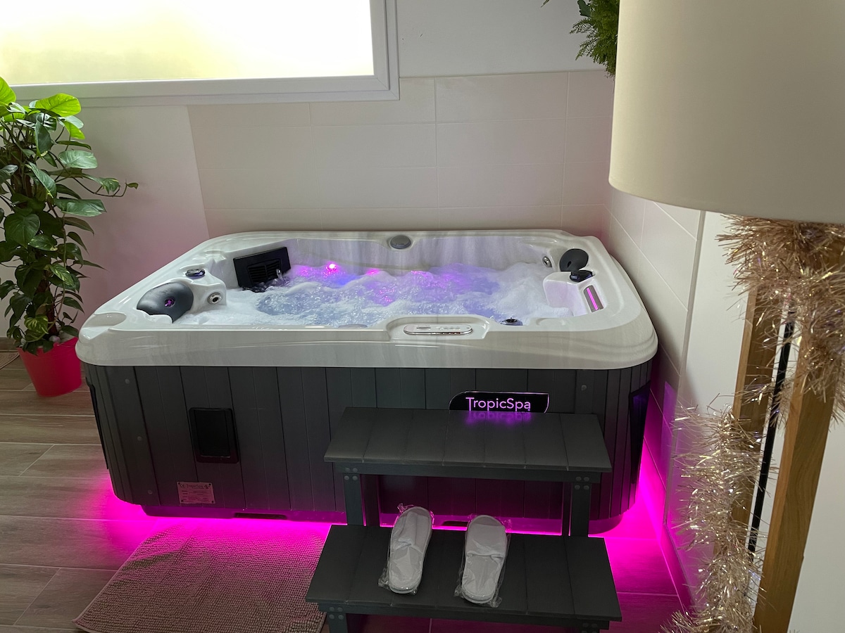 A private spa is featured in a bright space, surrounded by indoor plants. Soft lighting creates a relaxing ambiance. The spa is equipped with jets for comfort, and a small bench is placed nearby, alongside slippers, providing convenience for users.
