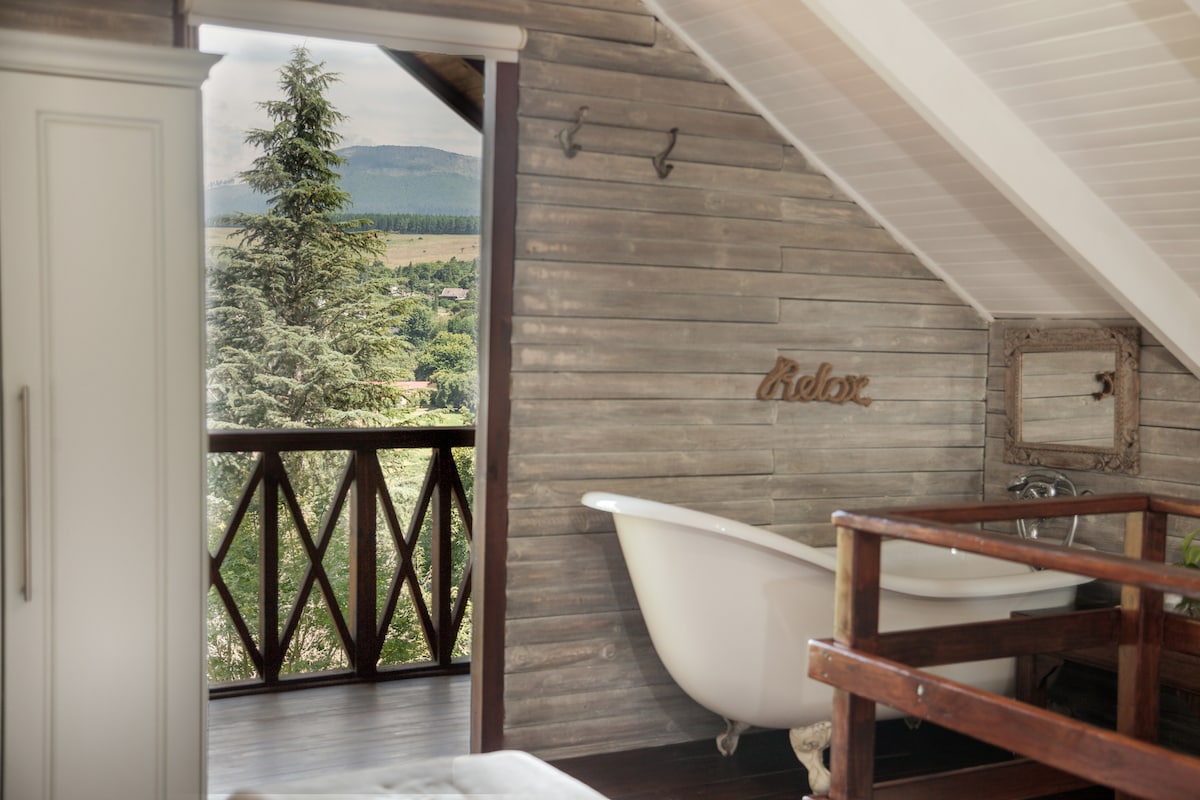 The interior showcases a charming freestanding bath positioned near large windows, revealing a view of the surrounding landscape. Natural wood paneling adds warmth, while a small balcony with wooden railings enhances access to the outdoors.