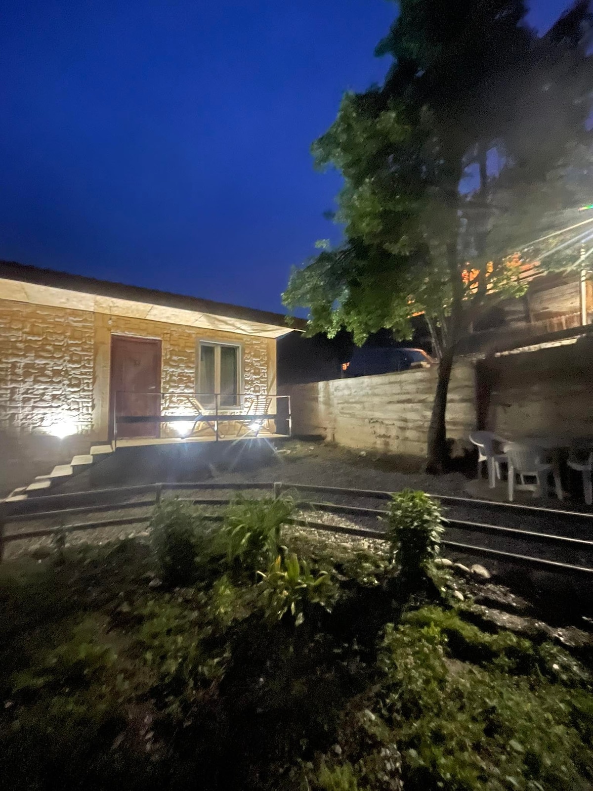 A rustic exterior of a guest house is displayed at night, with warm lights illuminating the staircase. The surrounding garden features a well-maintained pathway, greenery, and a seating area with several chairs, creating an inviting outdoor space.