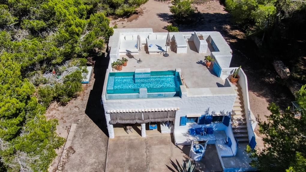 An aerial view of the chalet reveals a spacious terrace featuring a crystal-clear pool at the center. Surrounding areas include sun loungers and shaded spots, with lush greenery evident from the property. The structure is designed with bright white walls, blending harmoniously with its natural surroundings.