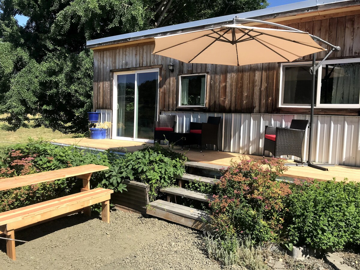 A welcoming outdoor deck features an umbrella providing shade over seating areas with chairs. Steps lead down to a landscaped garden with diverse greenery. The rustic guest house showcases wood paneling and large glass doors, inviting natural light into the space.