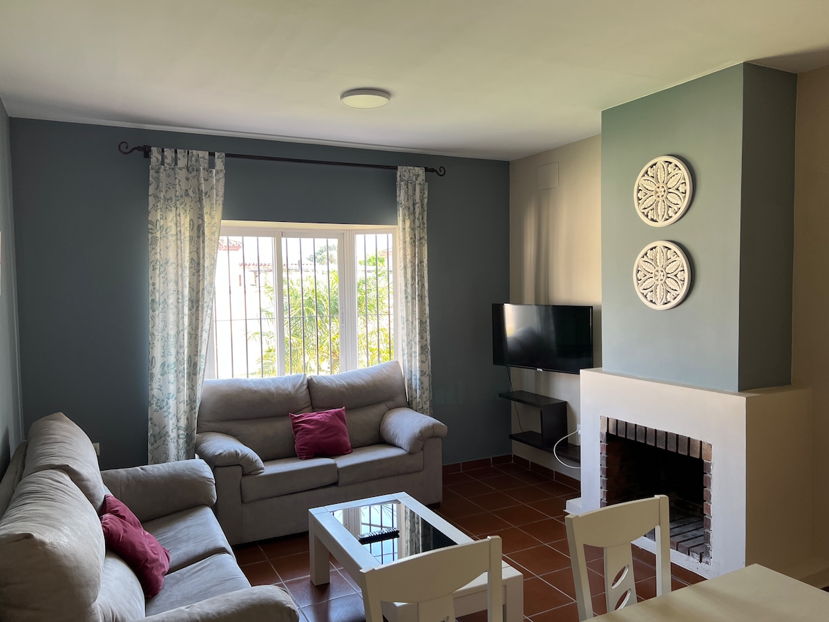 A living area features light-colored sofas with accent pillows, set around a glass coffee table. A television is positioned next to a decorative fireplace, while natural light enters through a window, framed by sheer curtains, illuminating the room's soft color palette.
