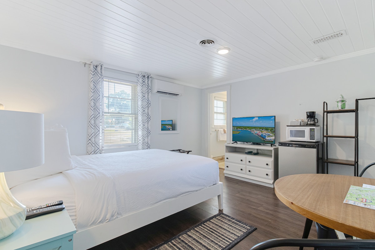 A bright room features a spacious bed with crisp white linens, alongside a flat-screen TV and a small dining table. A mini-fridge and microwave are neatly arranged on a shelf, with sunlight filtering through a window dressed with light curtains.