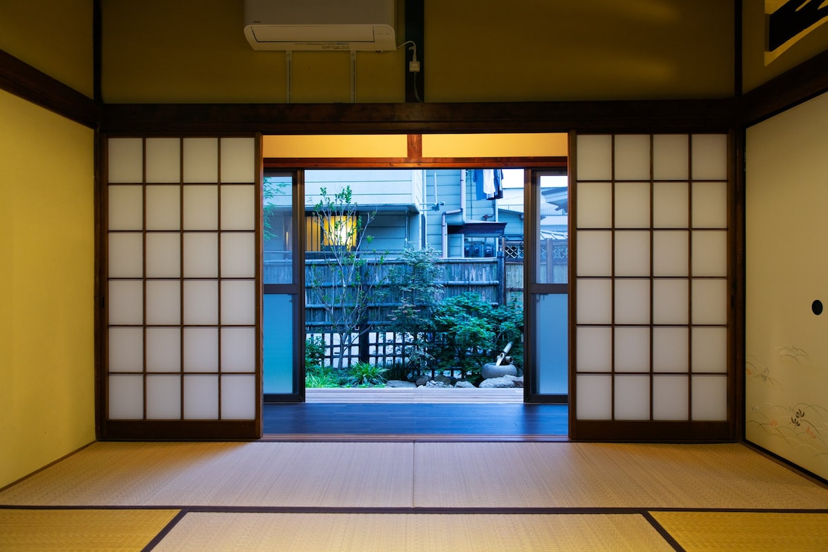 A traditional room features sliding shoji doors leading to a serene garden view. The space is adorned with tatami flooring and soft lighting, allowing for a peaceful atmosphere. Natural elements from the garden are visible beyond the open doors, enhancing the indoor-outdoor connection.