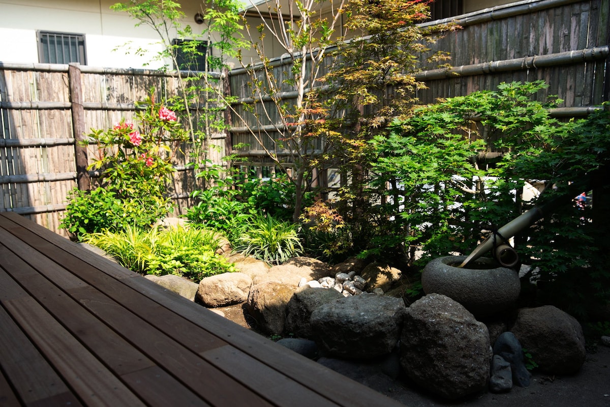 A serene garden area features a mix of greenery and flowering plants, bordered by wooden fencing. Natural stones are arranged artistically, and a small water feature is visible, contributing to a calm outdoor atmosphere. Sunlight filters through the leaves, casting gentle shadows.