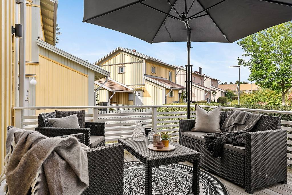 A covered outdoor seating area is furnished with two cozy chairs and a matching table. Plush throws are draped over the chairs, and a decorative rug lies underneath. Lush greenery and charming homes are visible in the background, creating a relaxed atmosphere.