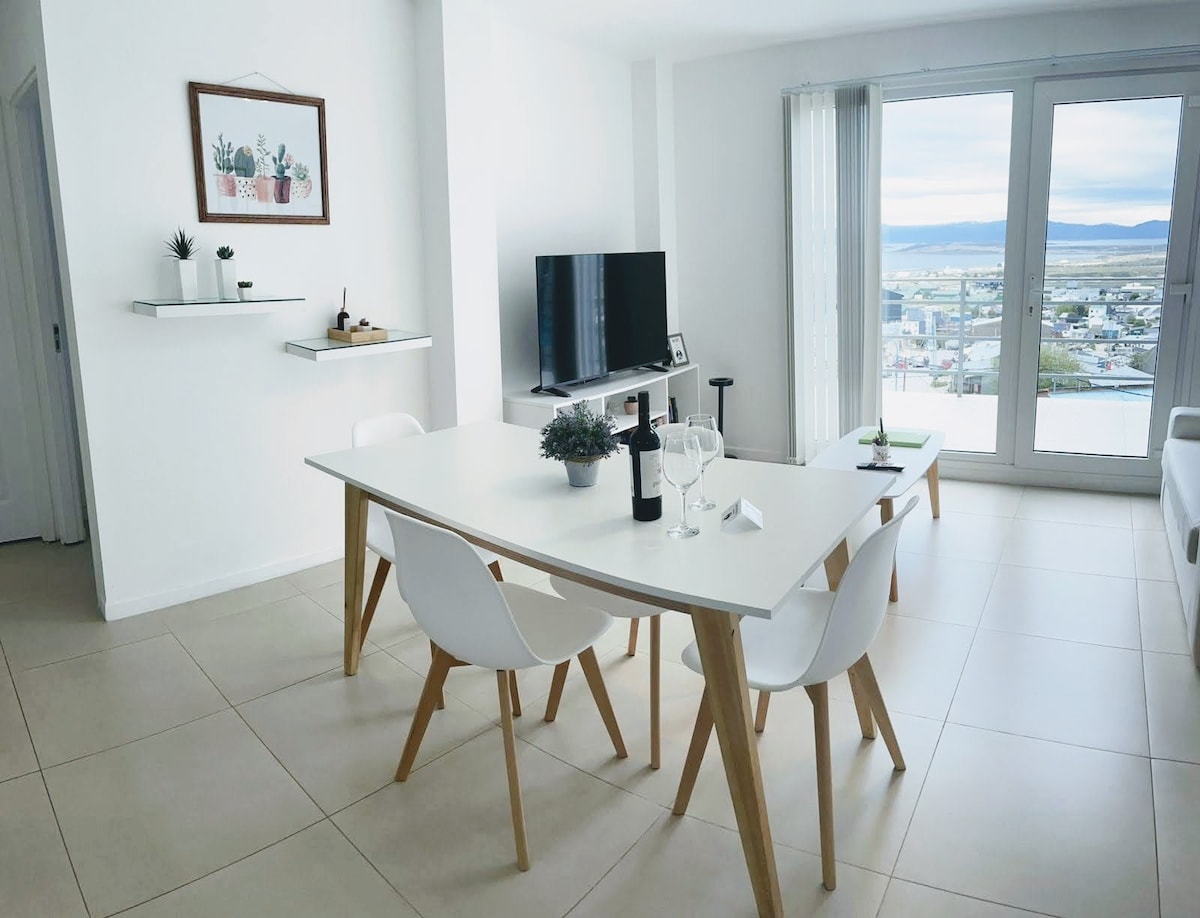 A bright living and dining area is shown, featuring a modern white table surrounded by four chairs. A television is mounted on the wall, and large windows provide views of the city and mountains in the background. Decorative elements include a plant and framed artwork.