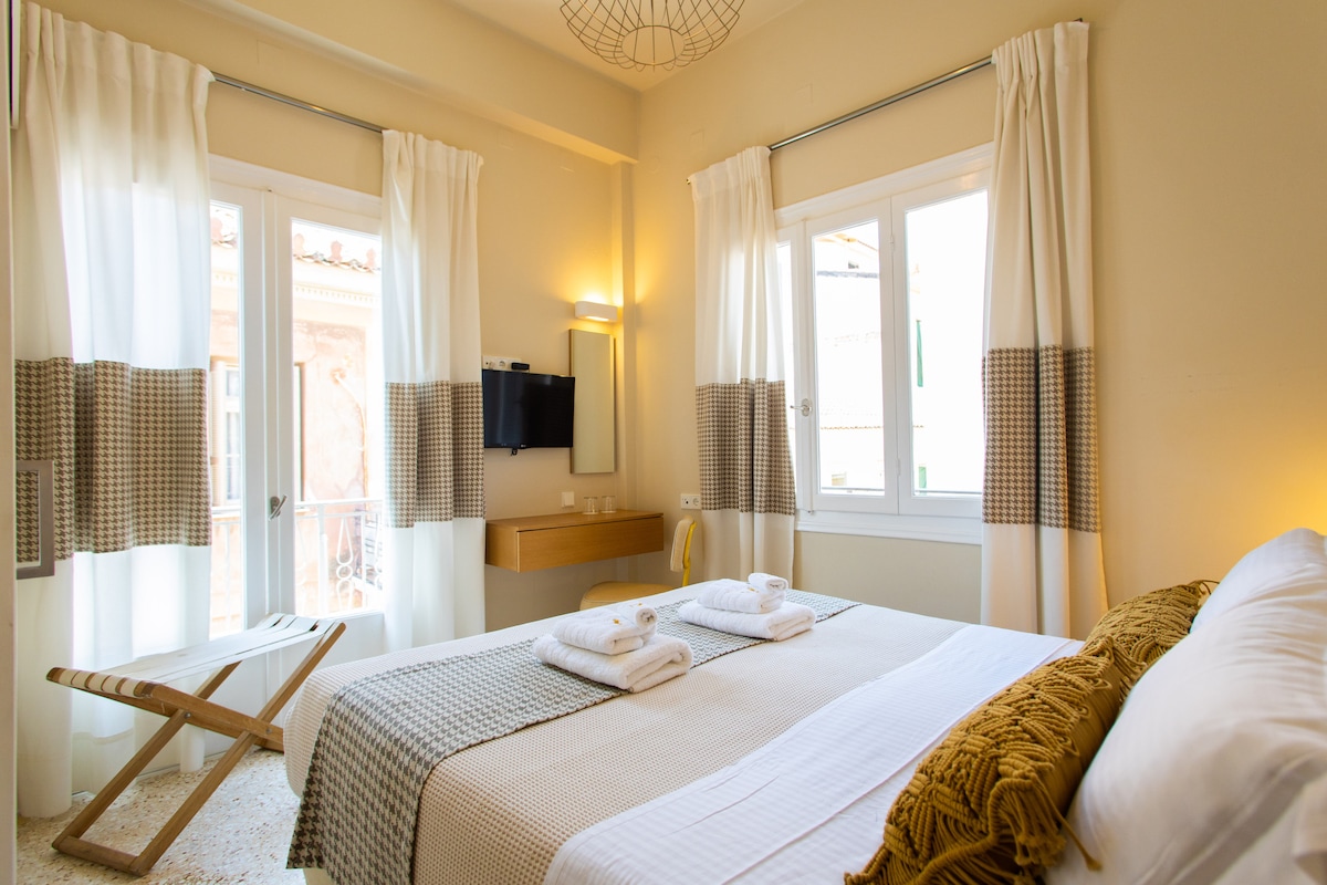 The room features a spacious bed adorned with neatly arranged towels. Natural light fills the space through large windows, complemented by soft curtains. A small desk and a flat-screen TV are positioned against the wall, enhancing the functional layout.