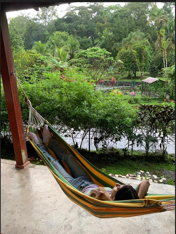A colorful hammock is suspended on a covered patio, with a guest reclining comfortably. Lush green vegetation surrounds the area, with vibrant flowers visible in the background. A peaceful river can be seen flowing among the greenery, creating a serene atmosphere.