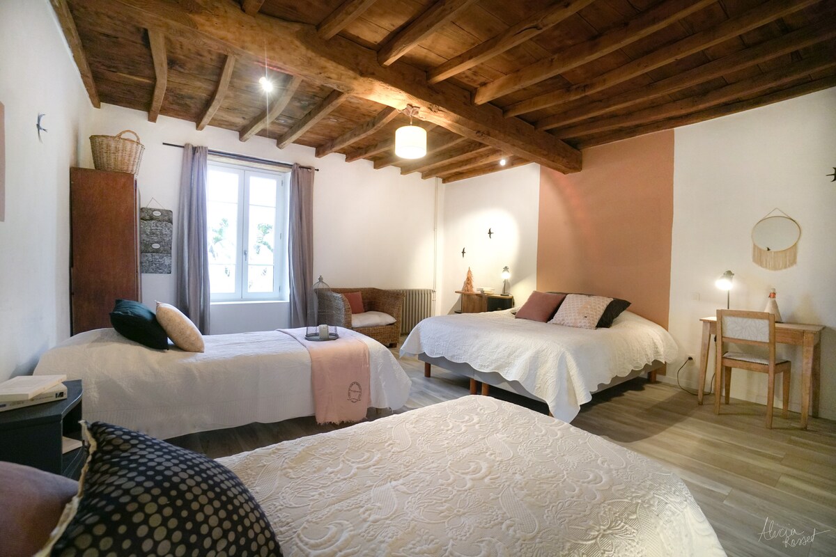 A spacious bedroom features three beds, including a double and two singles, all dressed in light linens. Exposed wooden beams adorn the ceiling, while a large window allows natural light to brighten the room. A small desk and a cozy seating area are also present.