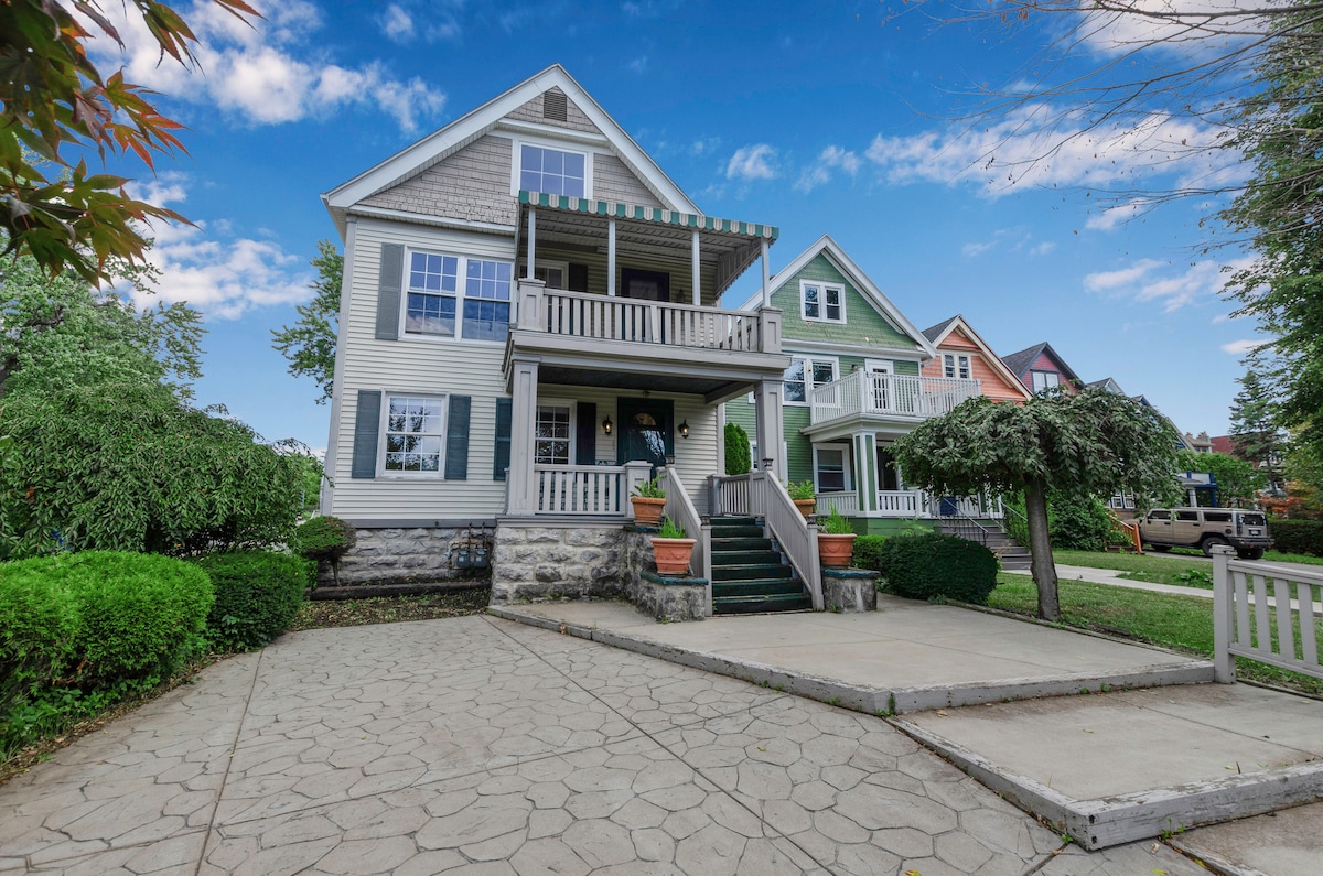 A charming exterior features a two-story house with a light grey façade and green accents. Two porches offer seating areas, surrounded by well-maintained shrubs and potted plants. A stone driveway leads to the entrance, complemented by neighboring colorful homes.