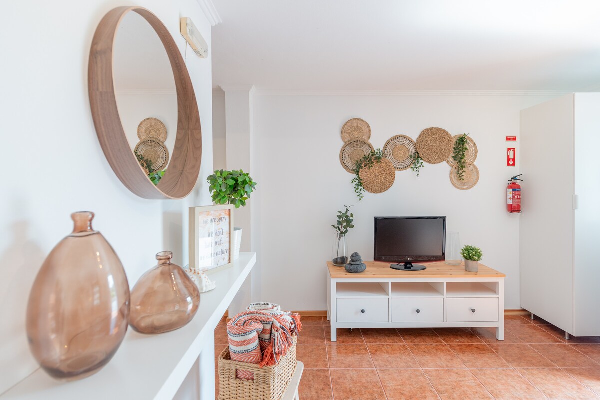 A welcoming living area is highlighted by a modern television on a light wood console. Decorative woven wall art pieces add texture to the space. A small potted plant and cozy throw blankets are positioned for easy access, enhancing the inviting feel of the room.