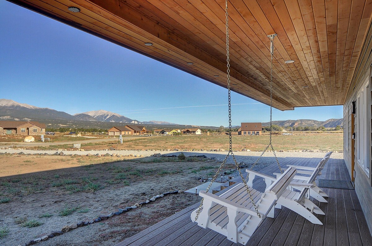 A large covered porch features a wooden ceiling and expansive views of the surrounding landscape. Comfortable swings provide seating, set against a backdrop of distant mountains and an open field. The porch looks out over a peaceful area with a few nearby homes.