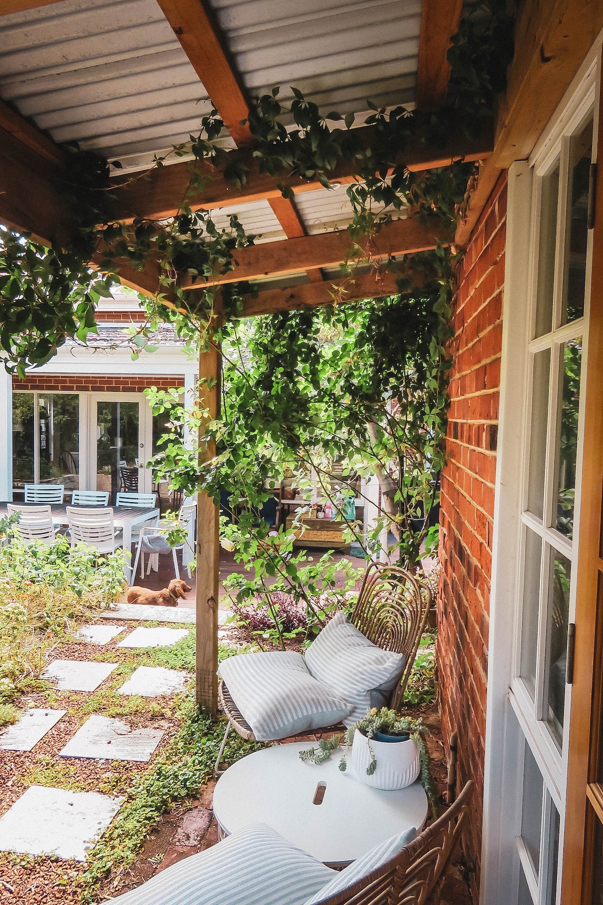 A charming outdoor seating area features two wicker chairs accompanied by a small white table. Lush greenery surrounds the space, with a pathway leading through a garden. Light filters through the leafy canopy above, creating a serene and inviting setting.