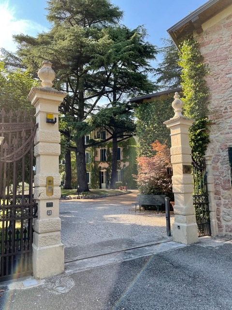 An entrance gate leads into a scenic property surrounded by mature trees and manicured landscaping. A gravel pathway extends towards a charming building, partially obscured by greenery. Sunlight filters through the trees, enhancing the serene atmosphere of the courtyard.