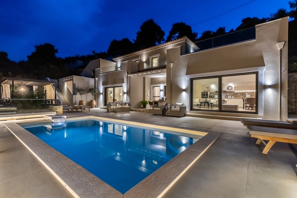 The modern villa is illuminated against the night sky, featuring a swimming pool surrounded by spacious outdoor seating. Large glass doors offer a view into the living area, enhancing the connection between indoor and outdoor spaces.